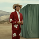 Steven Yeun in a cowboy outfit standing in front of a curtain-covered box.